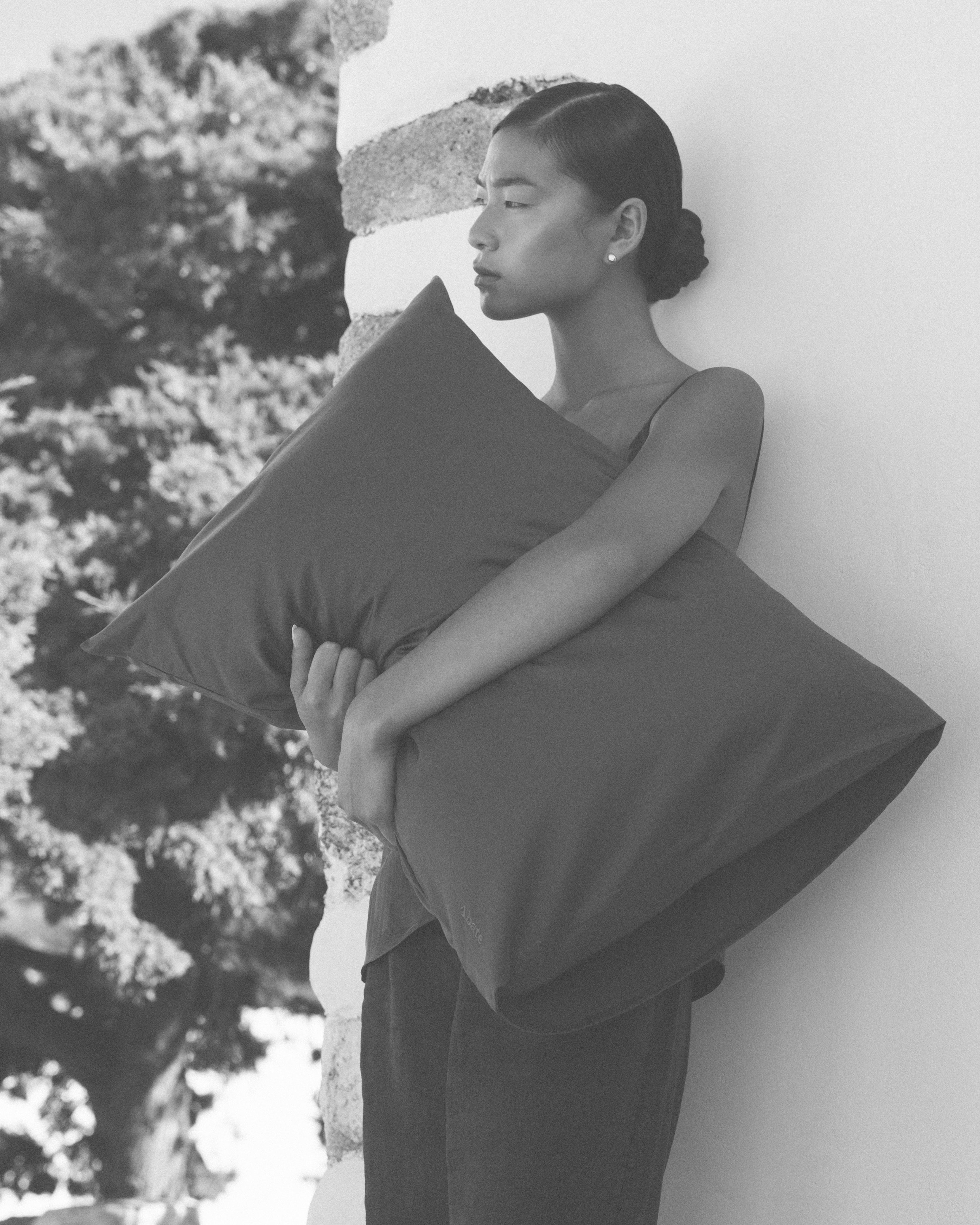 Woman holding a large pillow against a white wall with trees in the background
